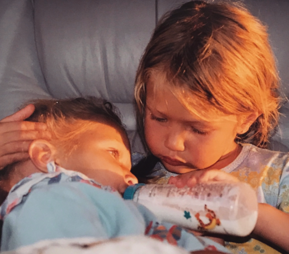 The elder sister Gigi is feeding the younger one Bella with a milk bottle